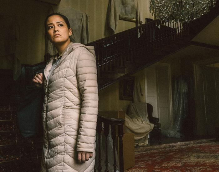 A woman wearing a coat stands in the dark, dusty hallway of a large house and is looking over her shoulder