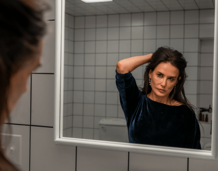 Demi Moore stands looking at herself in a bathroom mirror with smudged eye make-up