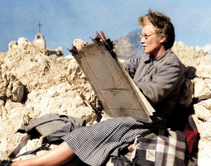 A woman sits sketching on a rocky beach