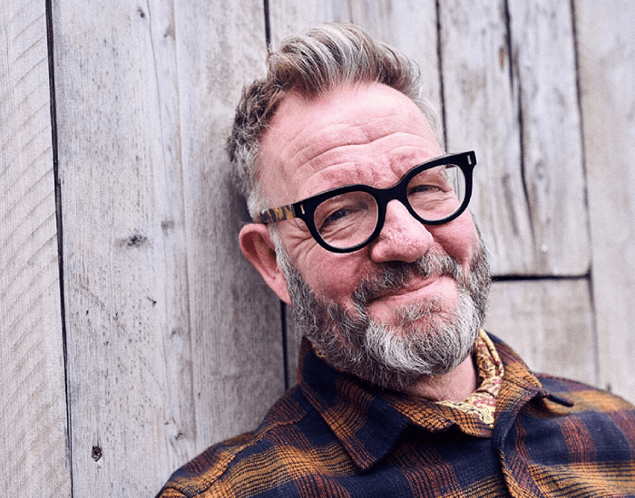 Pete Mckee with black glasses and tartan top leans against a wooden fence