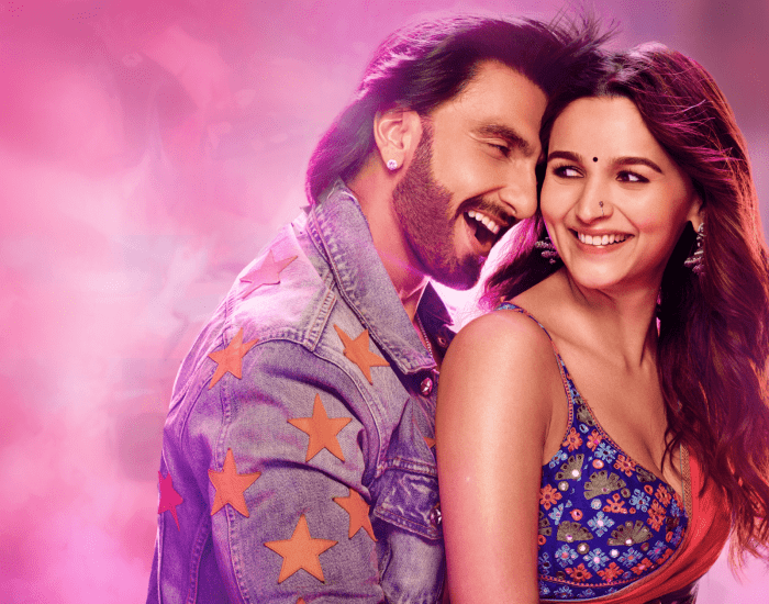 A joyful couple, a man and a woman, are embracing each other in a close-up shot against a vibrant pink, purple, and blue background. The man, on the left, has dark hair and a beard, and is looking towards the woman with a wide, happy smile. He is wearing a denim jacket adorned with orange star patches. The woman, on the right, has dark, wavy hair and is looking towards the man with an equally radiant smile. She is wearing a colorful, intricately patterned top and a sari. Both appear to be in their 30s.