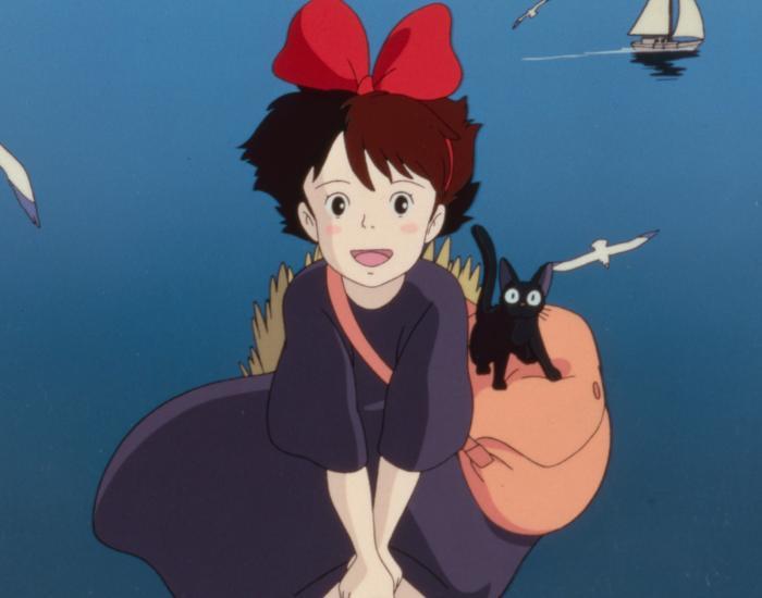 Kiki, a young girl with short brown hair and rosy cheeks, smiles brightly while flying. She wears her iconic oversized red bow and a loose-fitting dark purple dress. Riding on her large orange shoulder bag is her small black cat, Jiji, who has prominent, wide white eyes. They are flying over a vast, deep blue sea. In the upper right background, a small white sailboat floats on the water, accompanied by a few white seagulls flying nearby.