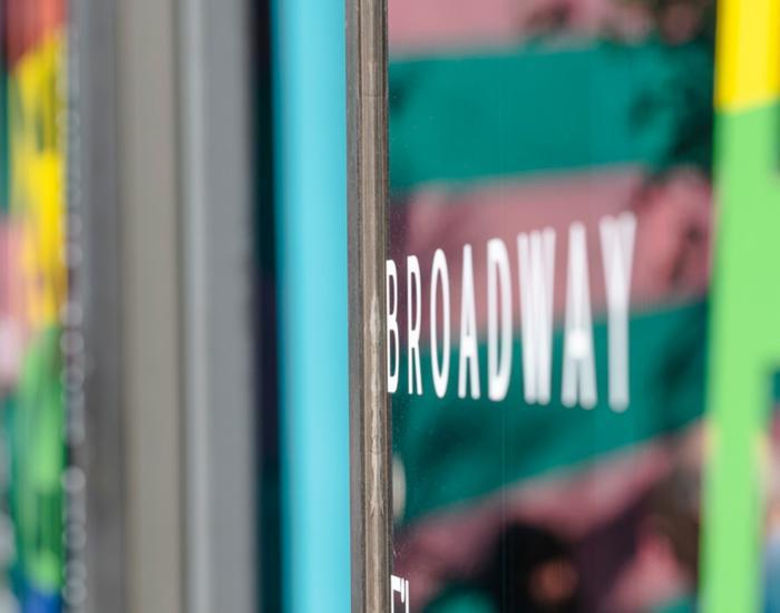 Image of Broadway Wordmark Logo emblazoned across the front of the building, The colourful reflection of the Broadway eye logo and rainbow colours in the foreground. 