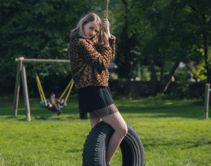 Beth ina leopard print jacket swings from a swing tyre in a park